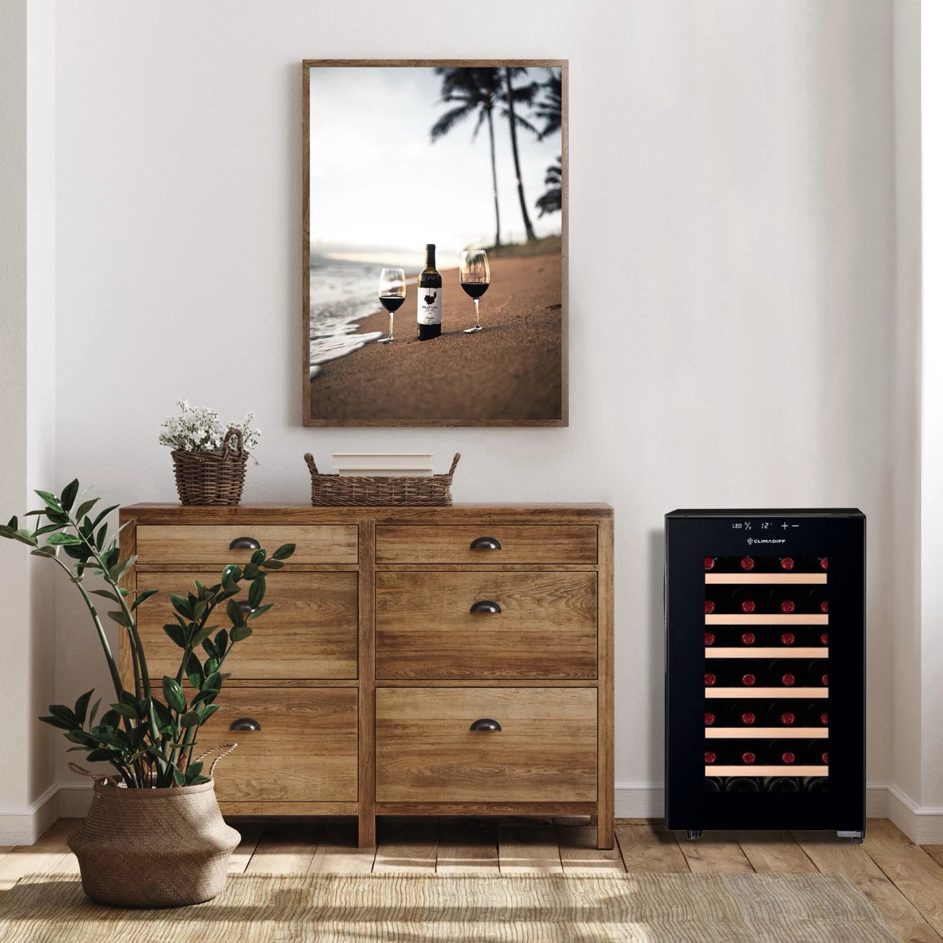 Der Weinkühlschrank Climadiff CUVEE28 mit schwarzer Glastür steht in einem stilvoll eingerichteten Raum neben einer Holzkommode. Durch die Tür sind mehrere Flaschen sichtbar, horizontal auf Holzregalen gelagert. Darüber hängt ein Strandfoto mit einer Weinflasche und zwei Gläsern.