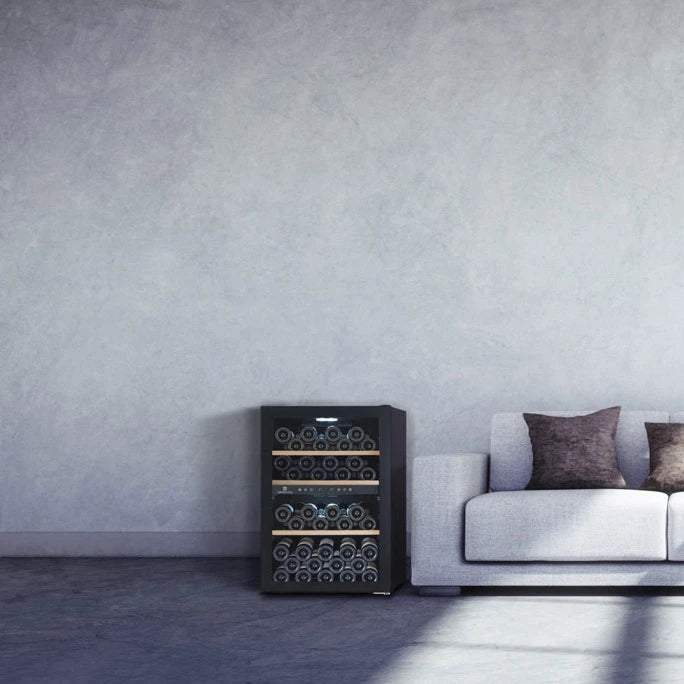Weinkühlschrank Cavecool Chill Topaz CC54DB-1, schwarz mit Glastür, gefüllt mit Weinflaschen auf Holzregalen, neben grauem Sofa vor Betonwand.