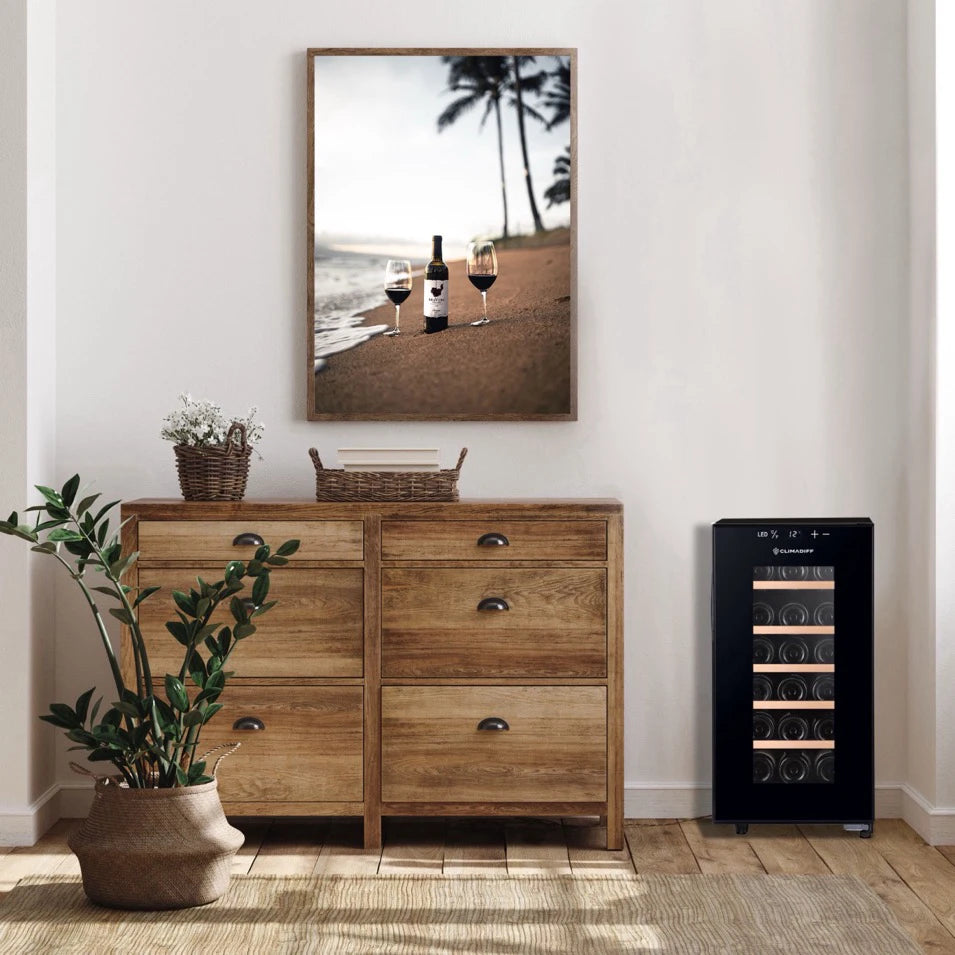Der Weinkühlschrank Climadiff CUVEE18 mit schwarzem Rahmen und Glastür steht neben einer rustikalen Holzkommode in einem hell eingerichteten Raum. Über der Kommode hängt ein gerahmtes Foto mit einer Weinflasche und zwei Gläsern am Strand, vor einer Kulisse mit Palmen und Meer.